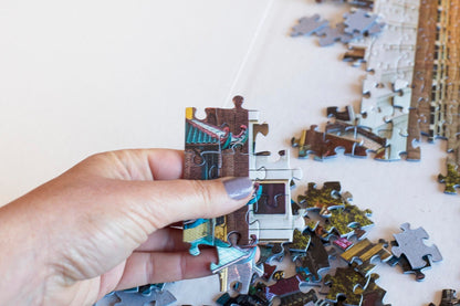 A hand holding a group of joined puzzle pieces that appear to a be a building with other pieces both joined and strewn in the background