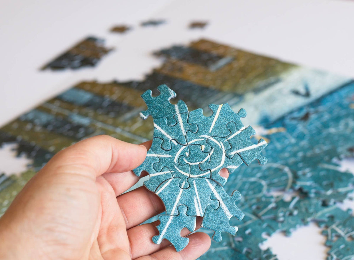 Hand holding a section of jigsaw puzzle featuring sunshine chalk art in front of a partially completed puzzle.
