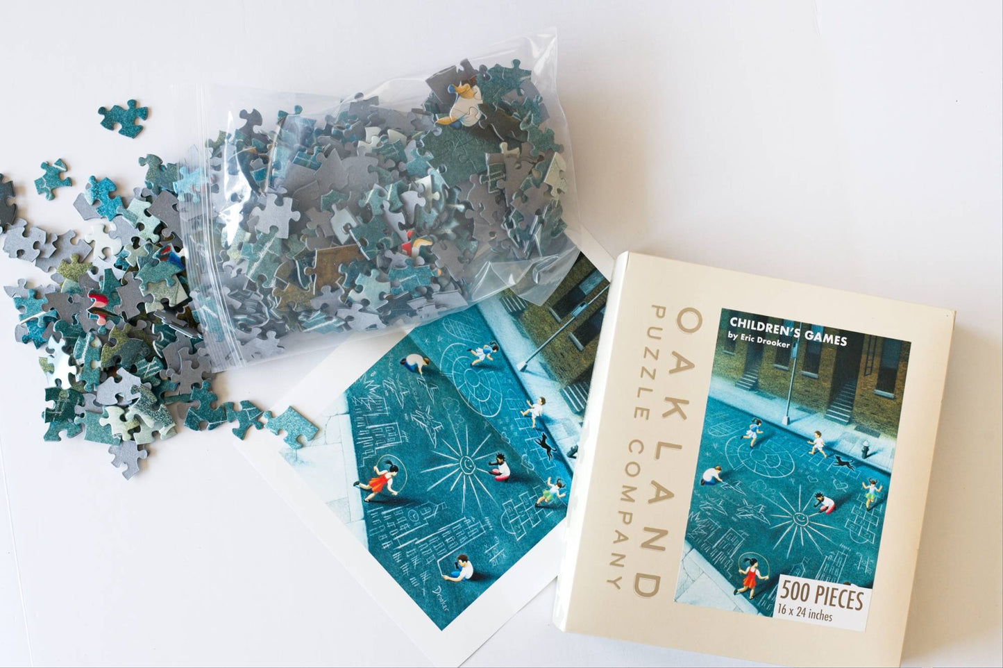 An open box with Oakland Puzzle Company branding with an image of Children Playing in front of Brownstones on the street with chalk next to a poster and resealable plastic bag spilling pieces