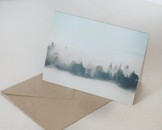 A photo of a greeting card with coniferous trees showing through wispy fog resting on a kraft brown envelope