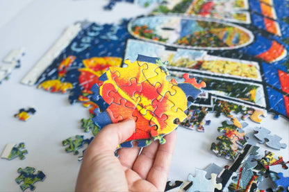 A hand holding a section of completed jigsaw puzzle featuring a California Poppy with a partially completed jigsaw puzzle in the background.