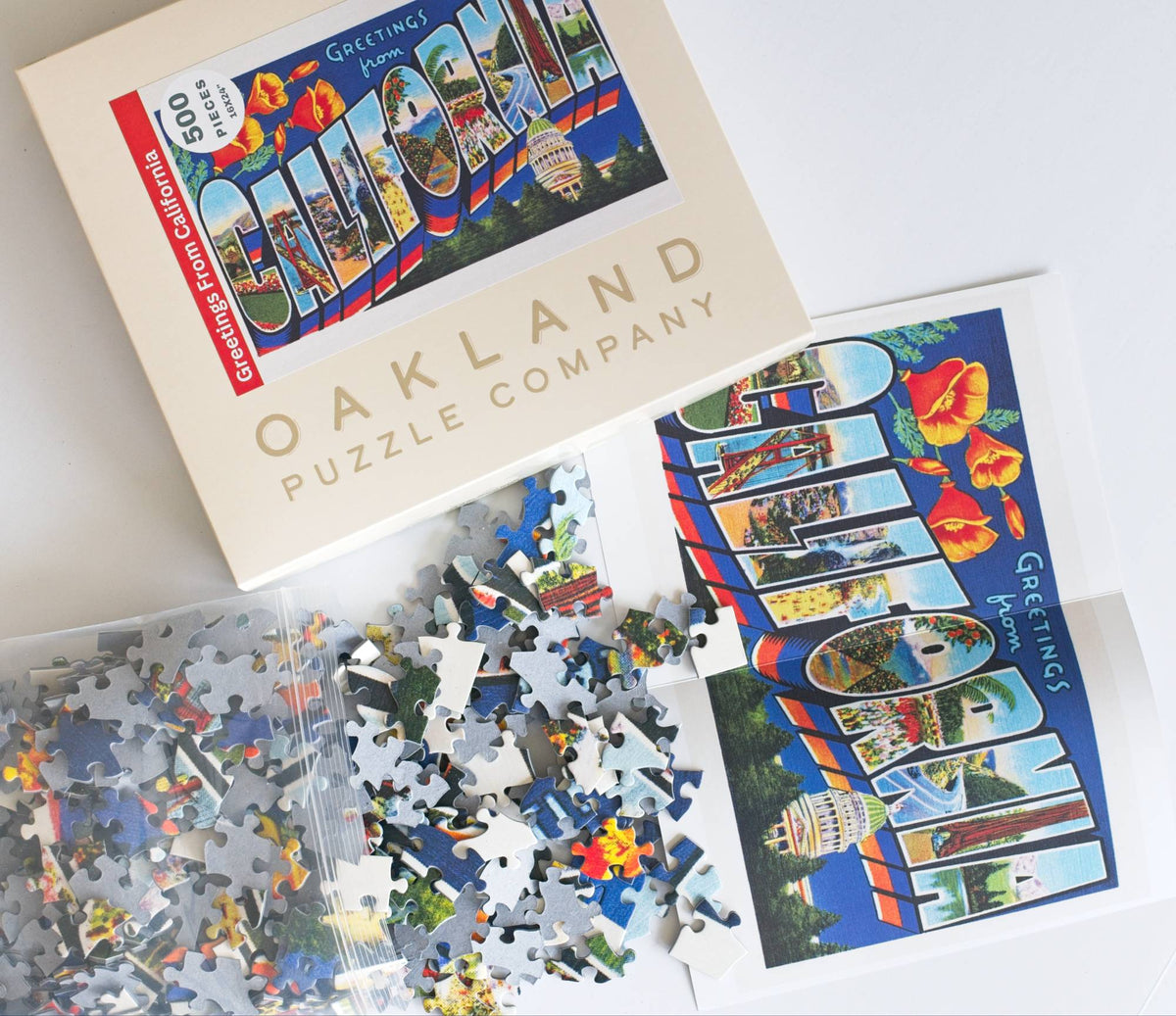 A resealable plastic bag spilling puzzle pieces out onto a reference poster showing a vintage California postcard iamge next to an open box with Oakland Puzzle Company branding. 