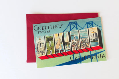 A photo of a vintage postcard with Greetings from Oakland printed in large letters filled with images resting on a red envelope on a white background
