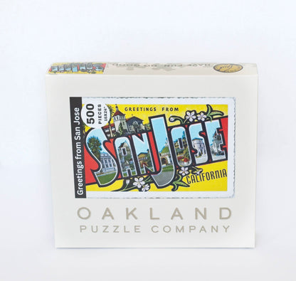 A box branded Oakland Puzzle Company labeled with a the image of a colorful vintage postcard reading Greetings from San Jose California 500 Pieces, 16x24"