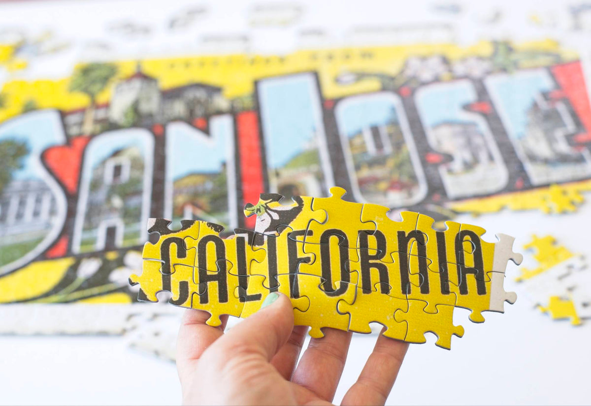 A hand holding interlocking puzzle pieces that show a yellow background with the word "California" in all caps in front of a slightly blurry partially completed jigsaw puzzle with the words San Jose.