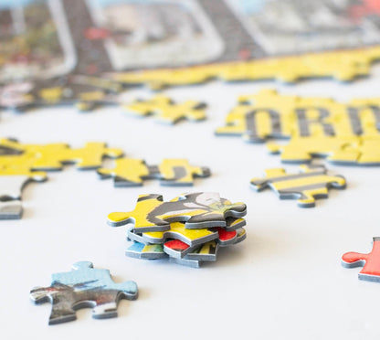 A close up of  a stack of colorful and sturdy jigsaw puzzle pieces made on blue board with a section of partially completed jigsaw puzzle in the background and scattered pieces around it.