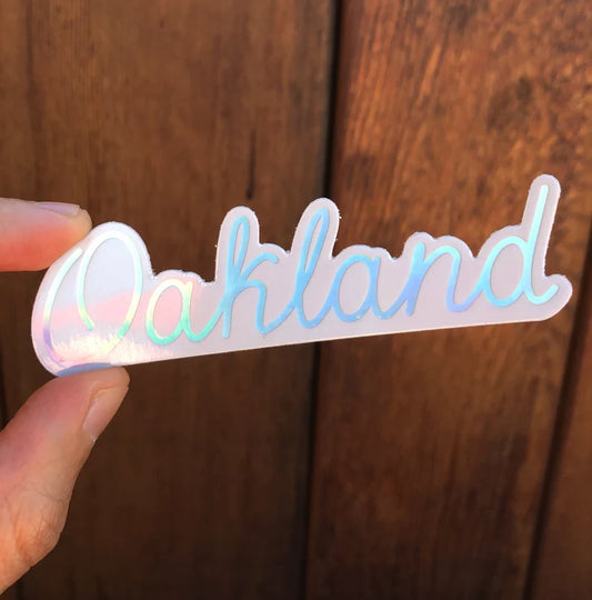 A blue and white holographic sticker with the name Oakland in cursive being held by a finger and thumb