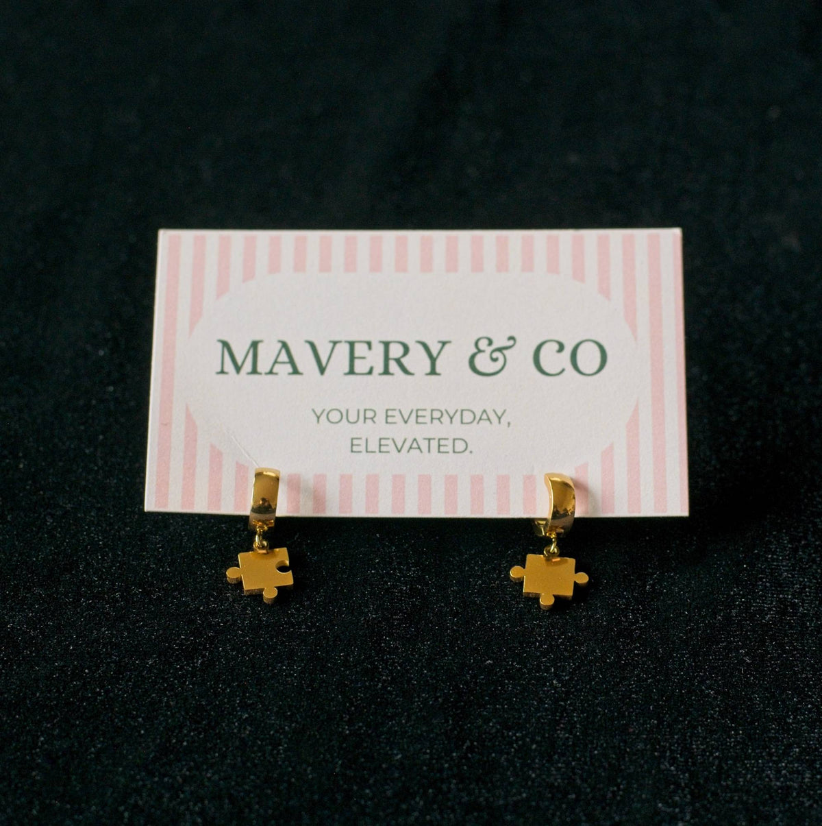 Gold small hoop earrings with dangling puzzle pieces on a Mavery & Co card against a black background