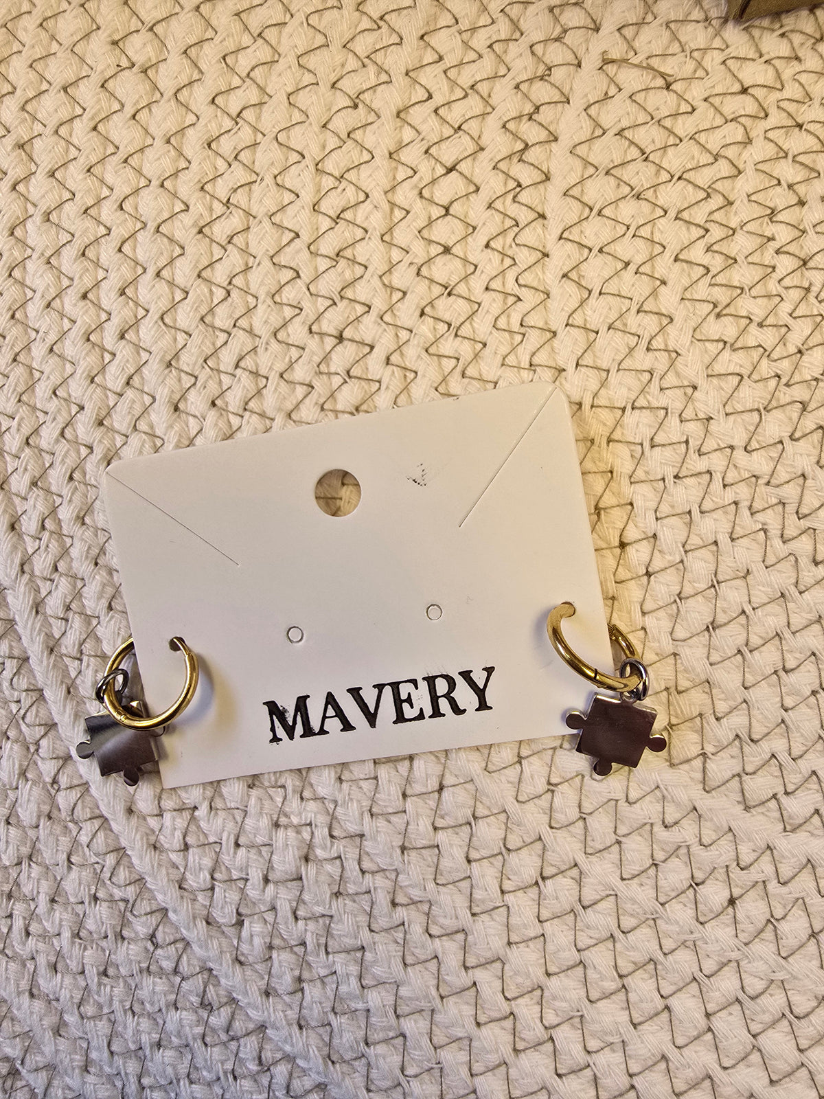 Pair of earrings with small gold hoops  and a dangling silver puzzle piece on a 'MAVERY' branded card against a textured beige background