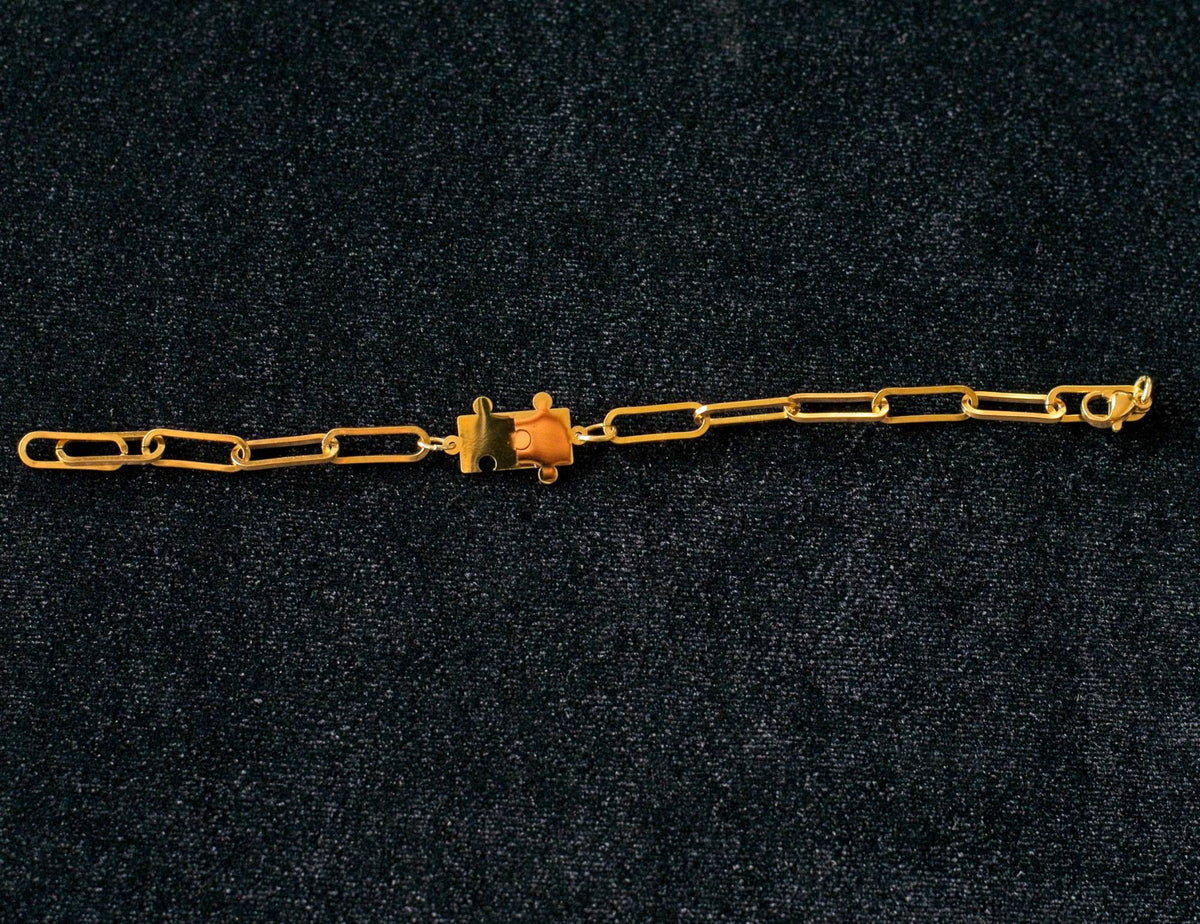 Gold paperclip chain bracelet with a puzzle piece connector charm on a dark textured background