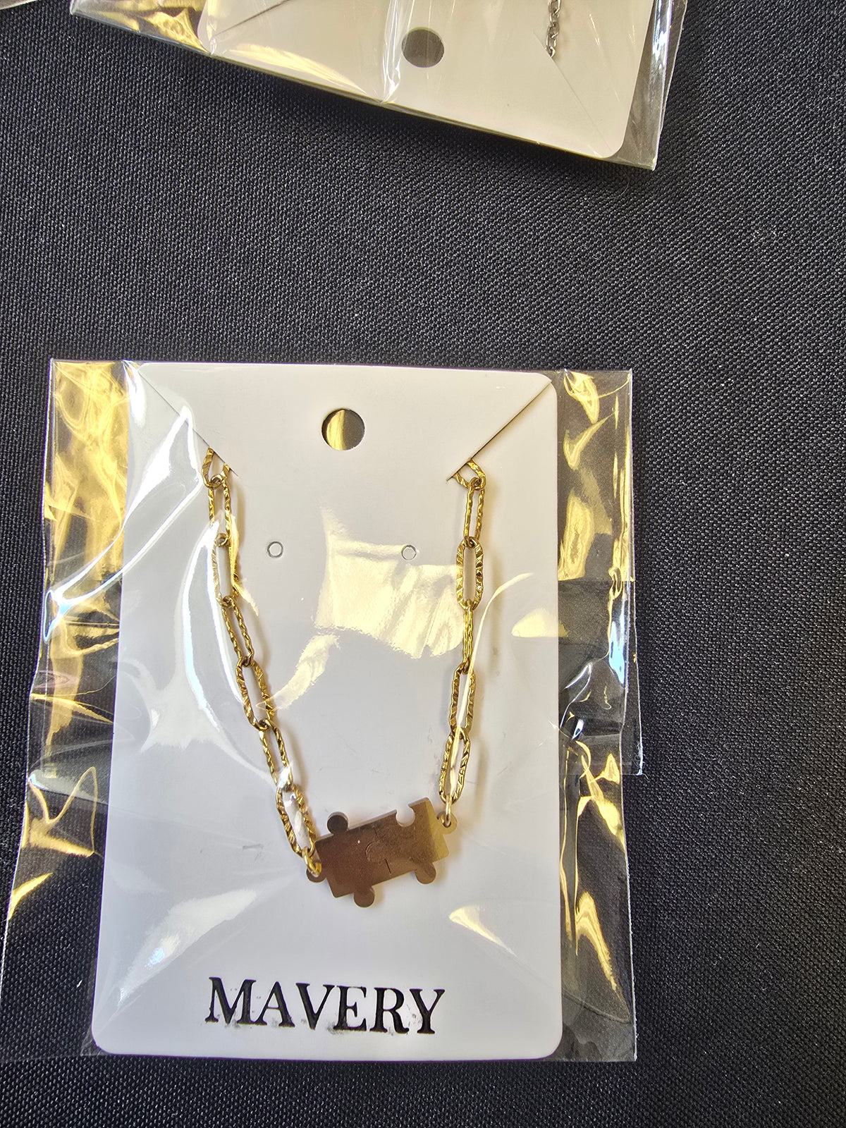 Gold  textured paperclip chain necklace with a puzzle piece connector charm on a 'MAVERY' branded card, against a dark fabric background.