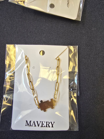 Gold  textured paperclip chain necklace with a puzzle piece connector charm on a 'MAVERY' branded card, against a dark fabric background.