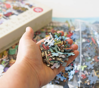 A hand holding puzzle pieces with an open puzzle box in the background