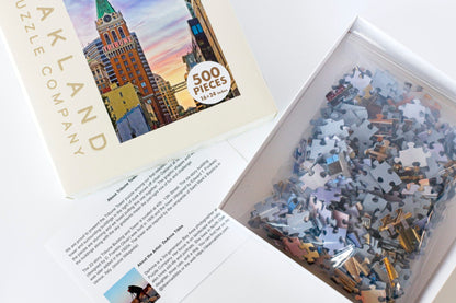 An open puzzle box with a puzzle guide. The box top is branded Oakland Puzzle Company and features the Tribune Tower image on the front. The box bottom has a plastic bag filled with puzzle pieces.