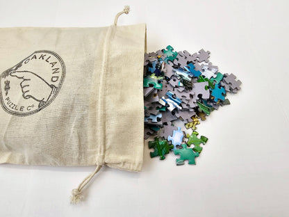 A cotton bag with the Oakland Puzzle Company logo with bright green, blue, and yellow puzzle pieces spilling out.